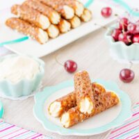 Three gluten free brandy snaps stacked on a plate, with a bowl of fresh cherries in the background.