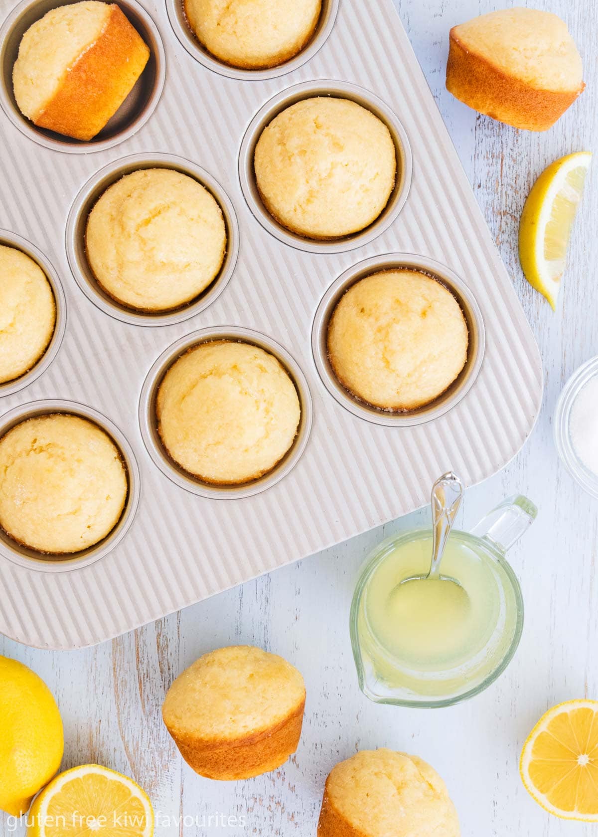 GF crunchy lemon muffins in a silver muffin tin, with a small glass jug of syrup, and sliced lemons on a blue background.