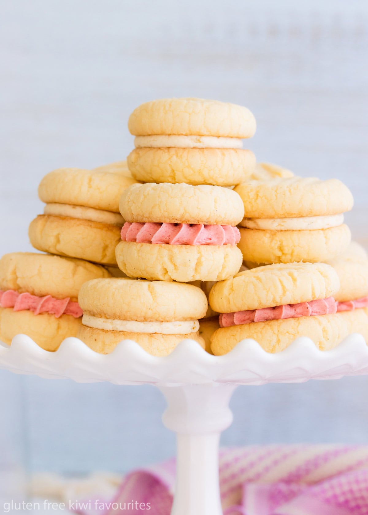 Gluten Free melting moment biscuits filled with pink and white buttercream on a white milk glass cake stand on a pale blue background.
