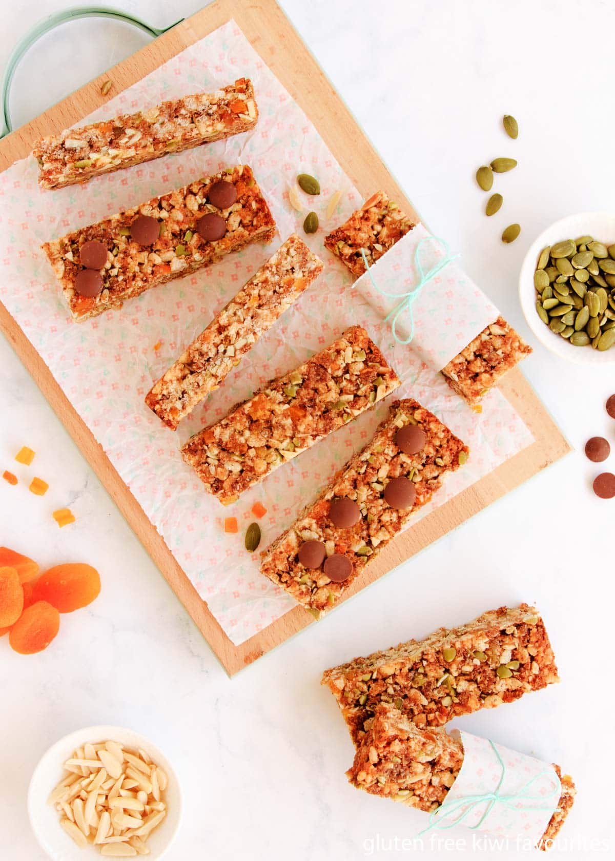 Muesli bars on a wooden board - some are topped with chocolate chips, and two are wrapped with baking paper and twine.