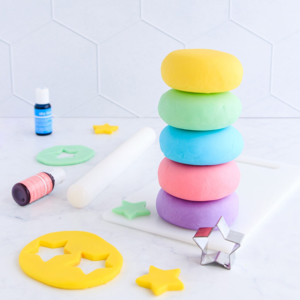 Five stacked balls of playdough - yellow, green, blue, pink and purple on a grey background, with yellow play dough star cutouts and a star cutter.