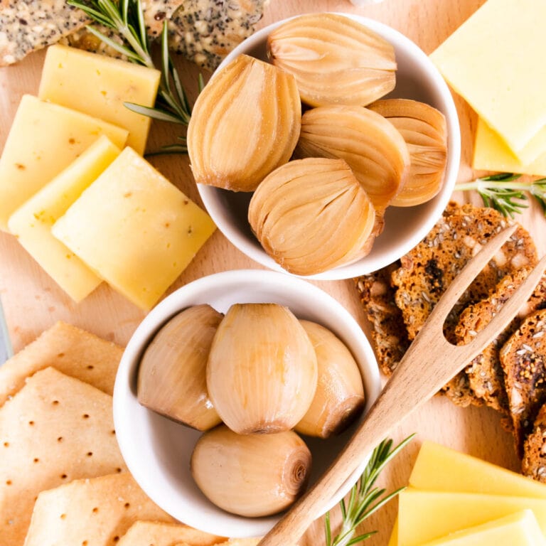 Close up of a cheese board featuring two small dishes of gluten free pickled onions, with a small wooden pickle fork, cheese and crackers.