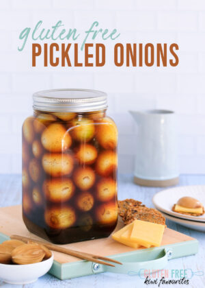 Gluten free pickled onions in a large glass jar, sitting on a wooden cutting board with cheese and crackers, and a small dish of halved pickled onions.