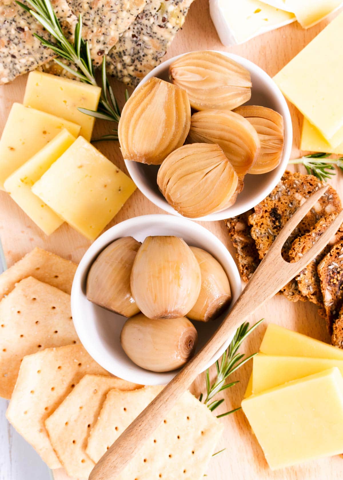 Close up of a cheese board featuring two small dishes of gluten free pickled onions, with a small wooden pickle fork, cheese, crackers and sprigs of rosemary.
