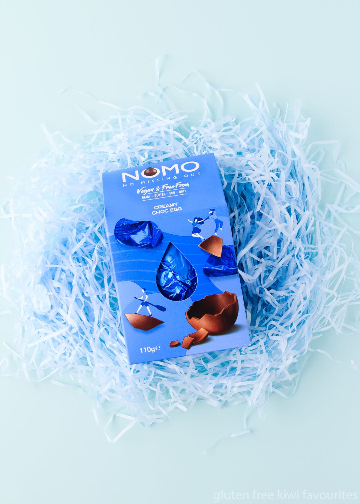 A NOMO gluten free, dairy free, nut free, vegan easter egg sitting on a nest of pale blue shredded paper.