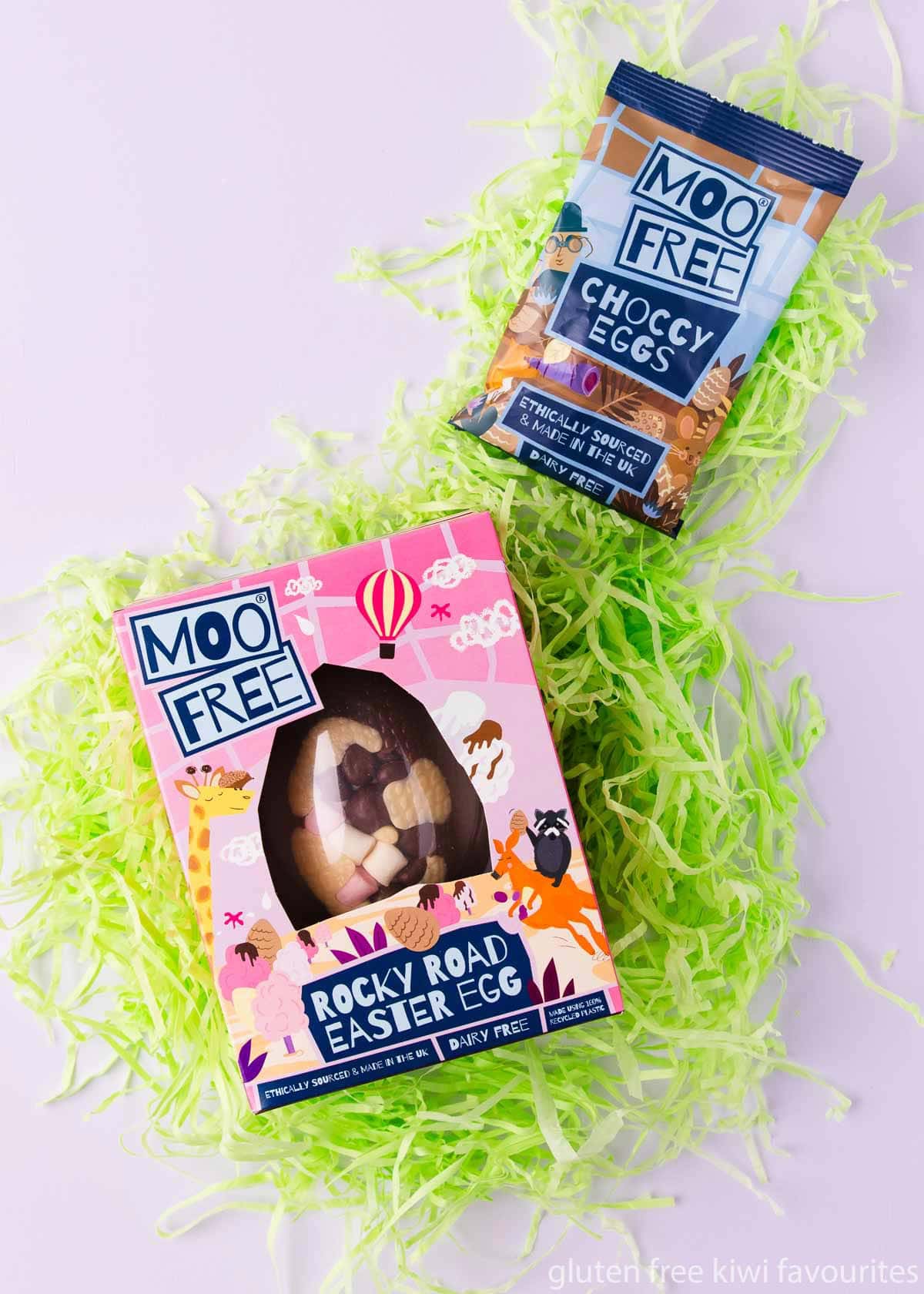 A Moo Free rocky road Easter egg in a colourful box, and a small blue bag of Moo Free mini choccy eggs, on green shredded paper, on a light purple background.