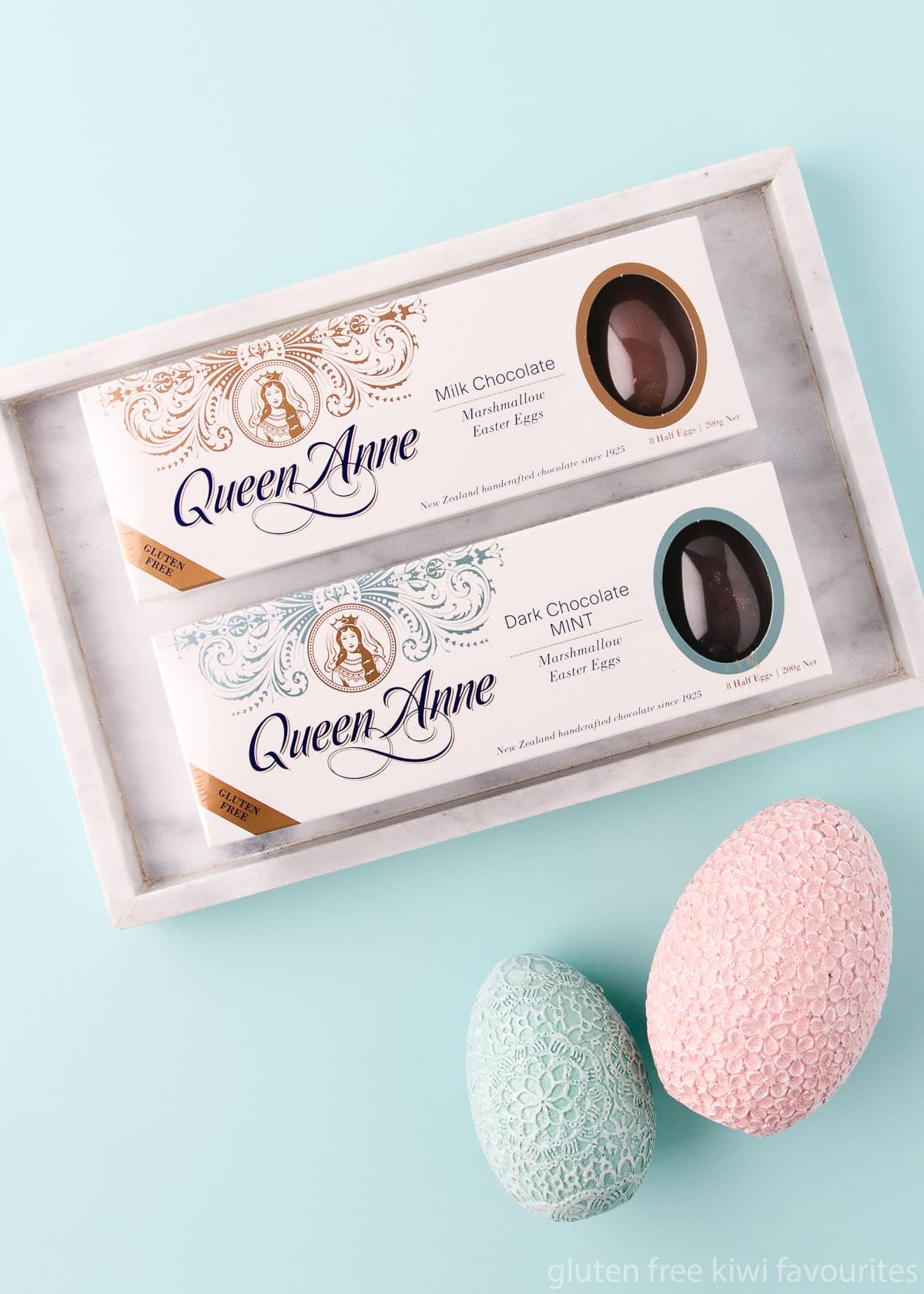 A box of gluten free Queen Anne milk chocolate marshmallow eggs and dark chocolate mint marshmallow eggs in a marble tray on a mint green background.