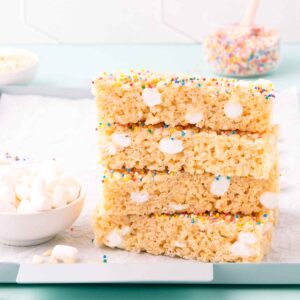 Four stacked pieces of gluten free rice bubble marshmallow slice on a light blue metal tray, with scattered mini marshmallows.