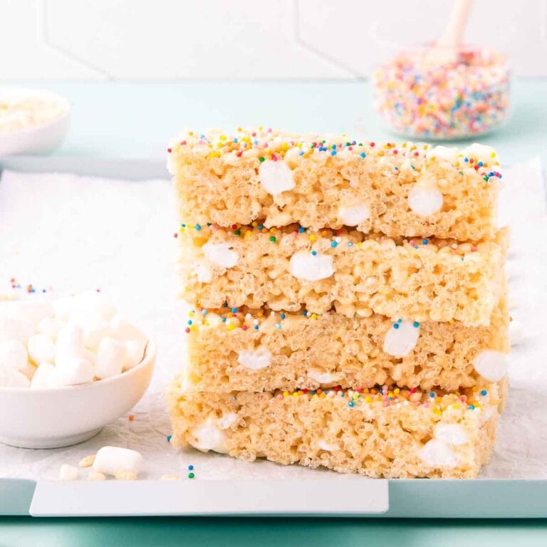 Four stacked pieces of gluten free rice bubble marshmallow slice on a light blue metal tray, with scattered mini marshmallows.