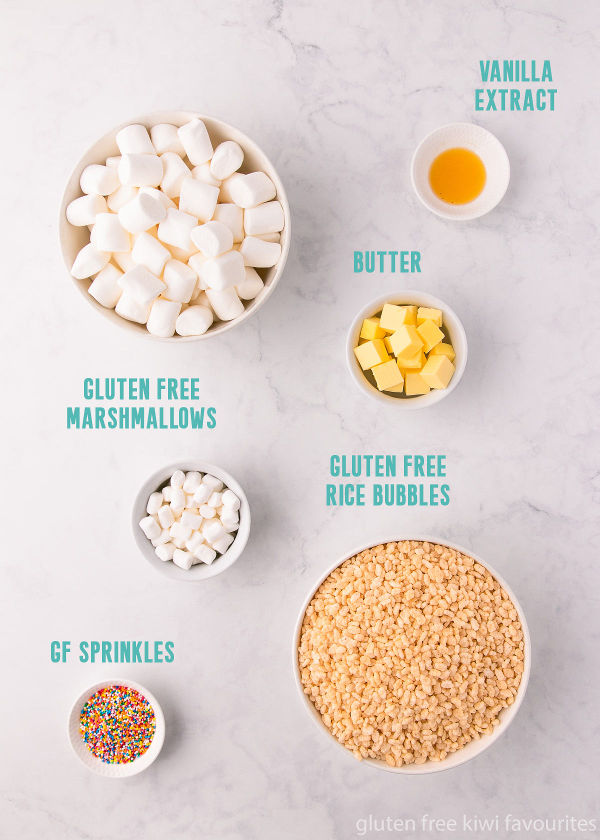 The slice ingredients - marshmallows, gluten free rice bubbles, butter, vanilla and sprinkles, in white bowls on a grey marble background. 