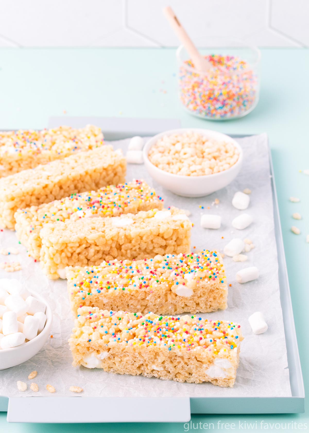 Six pieces of slice arranged in a curved line on a light blue metal tray, with small bowls of marshmallows, rice bubbles and rainbow sprinkles.