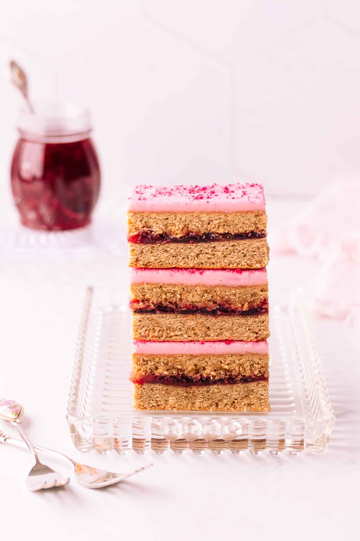 Three stacked pieces of Belgian slice on a rectangular glass plate, with a small jar of raspberry jam in the background and two cake forks in the foreground.