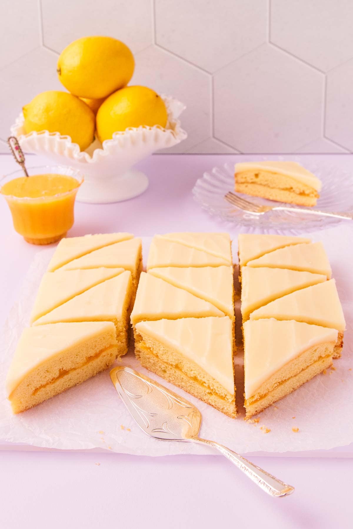 The pieces of lemon slice on a sheet of crinkled baking paper, with a silver cake server, a small glass dish of lemon curd and a bowl of lemons in the background.