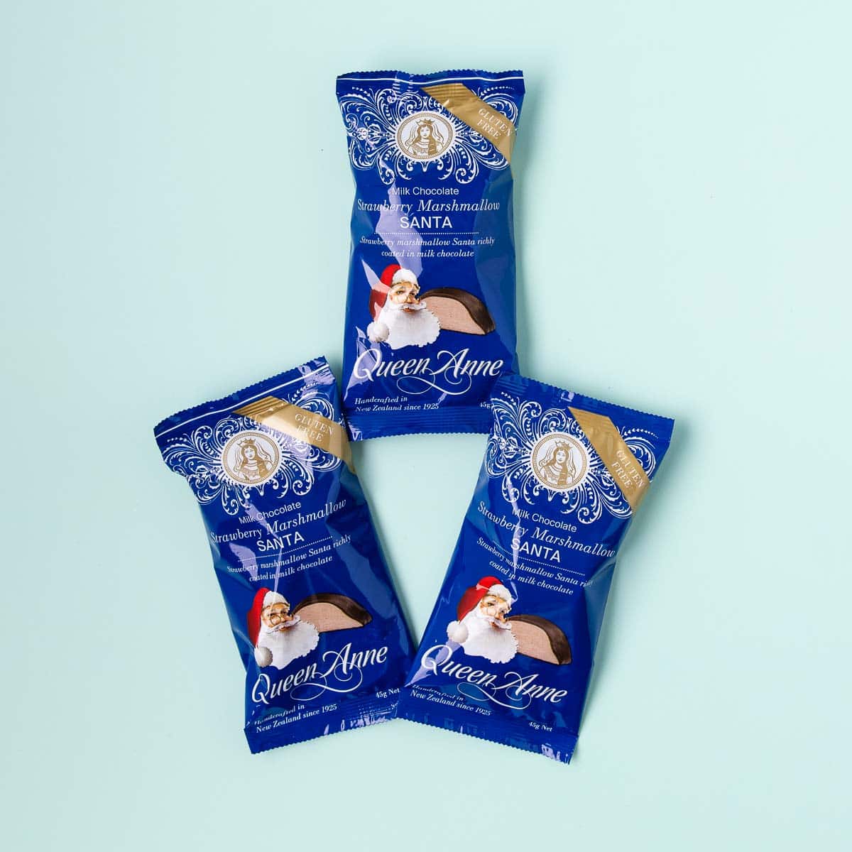 Three Queen Anne marshmallow Santas in blue plastic packaging, on a mint green background.