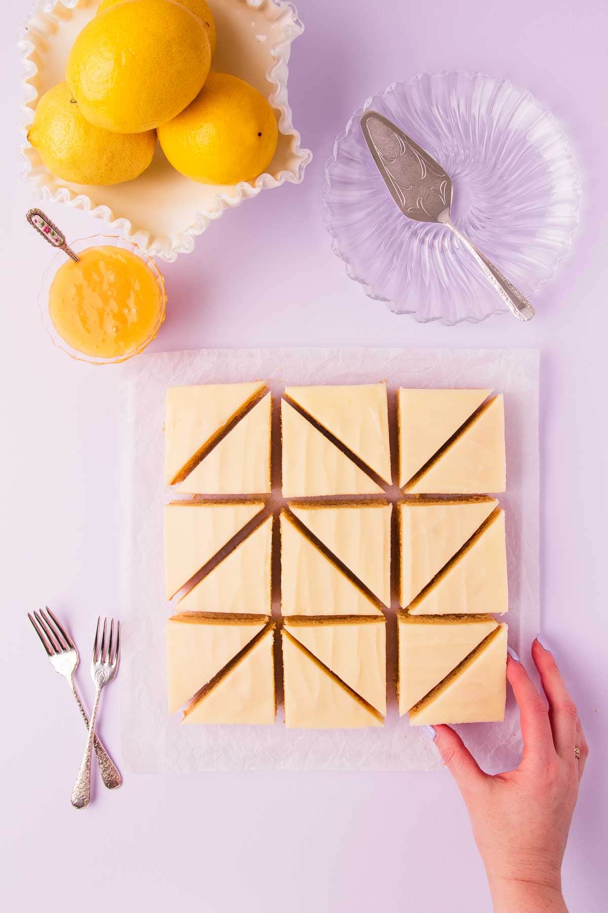 Lemon slice that has been cut into 16 triangle shaped pieces, on a light purple background, with a bowl of lemons, glass plates and cake forks. 