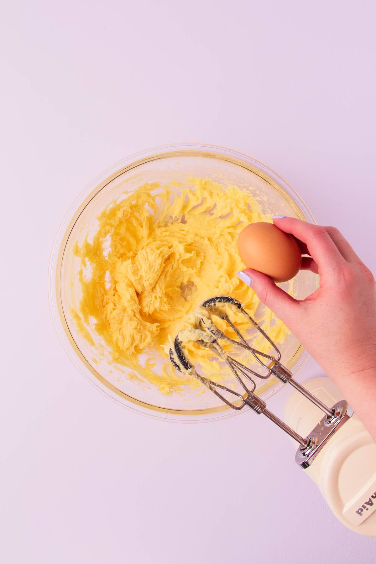 A glass bowl with creamed butter and sugar, and a hand about to crack and egg into the bowl.