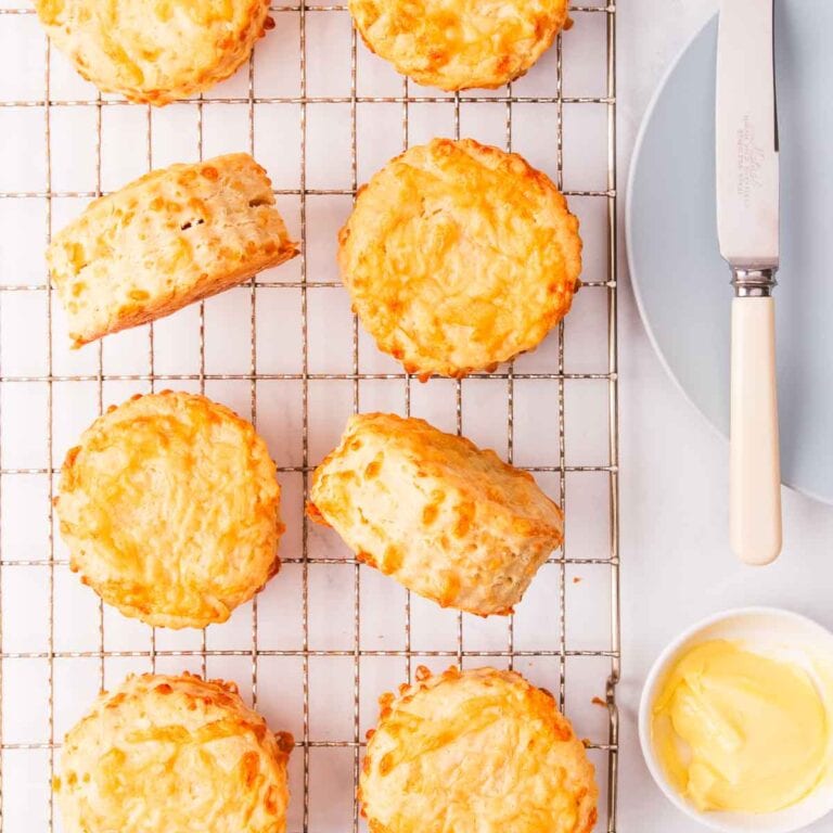 8 cheese scones on a wire cooling rack, with a small dish of butter and a light blue plate with a bone handled knife resting on it.