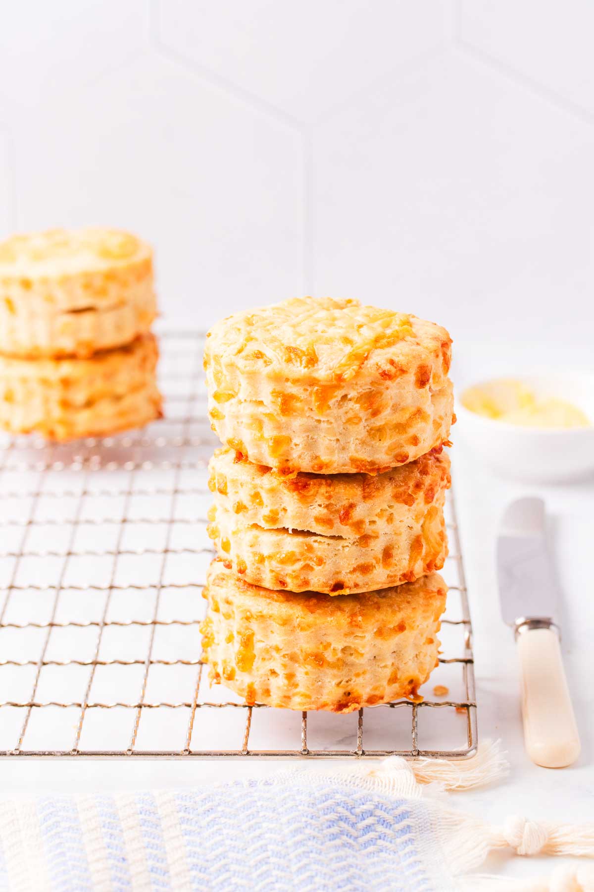 A stack of three cheese scones on a wire rack against a grey tiled background, with a small dish of butter and a bone handled knife.
