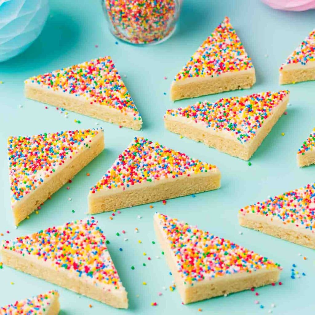Triangular pieces of fairy bread slice - a light coloured slice topped with buttercream and rainbow sprinkles - on a mint green background.