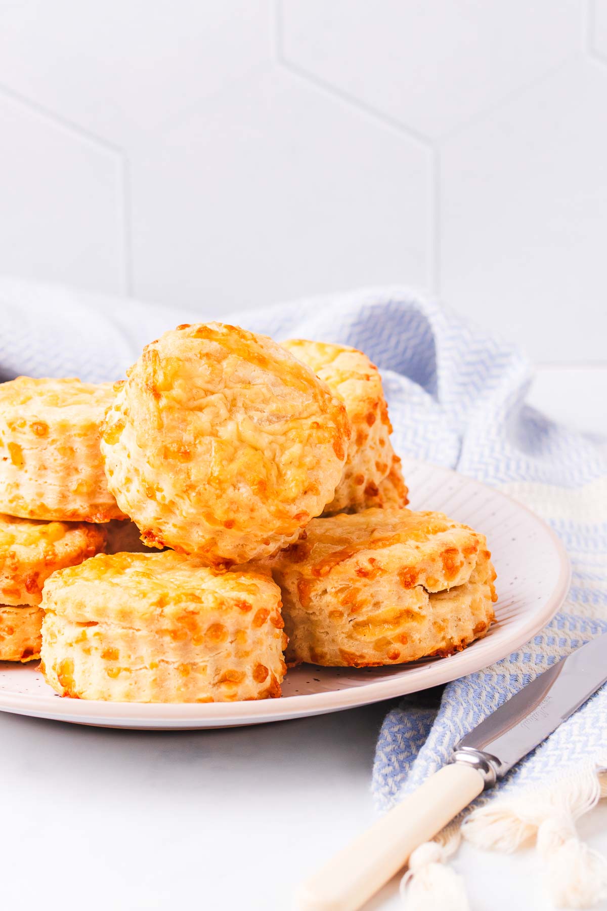 Gluten free cheese scones piled onto a large plate, with a pale blue tea towel and a bone handled knife.