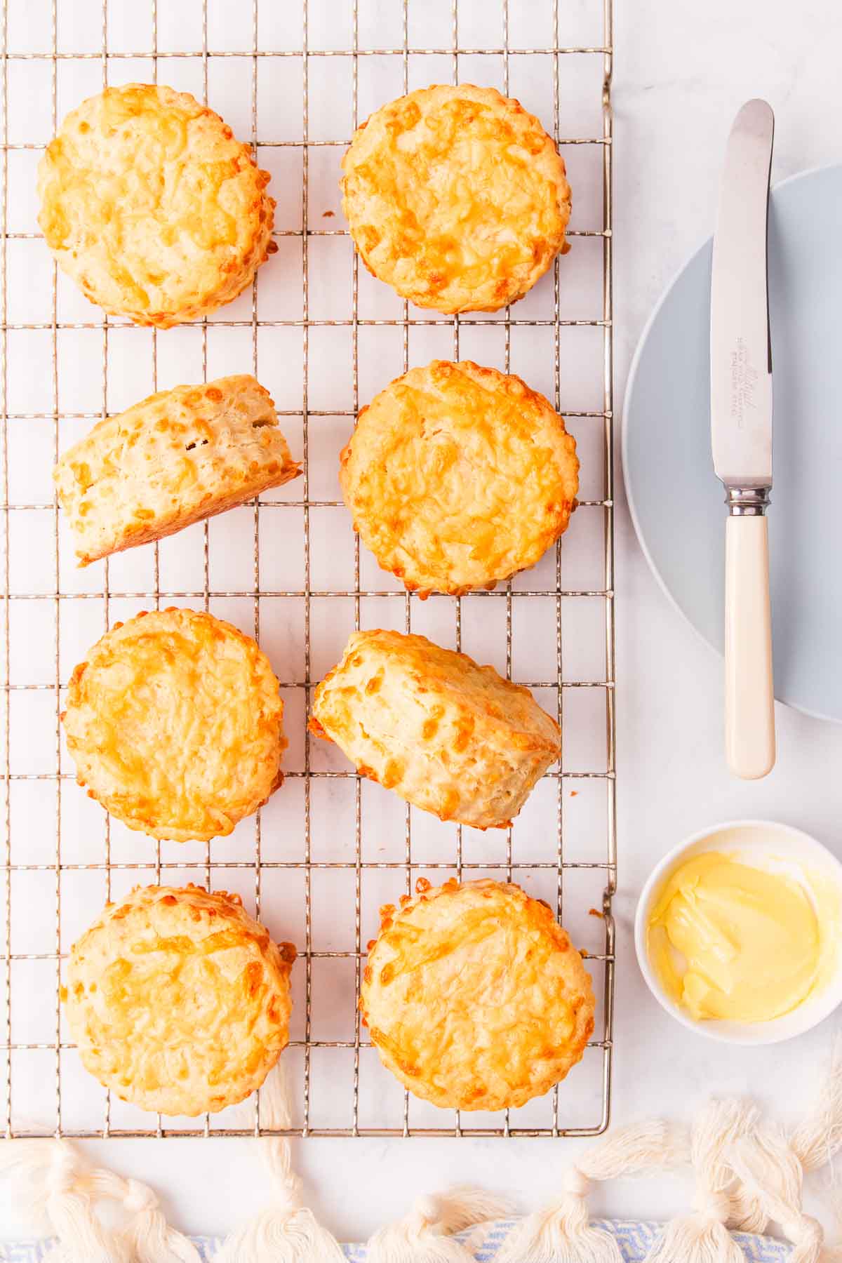 8 cheese scones on a wire cooling rack, with a small dish of butter and a light blue plate with a bone handled knife resting on it.
