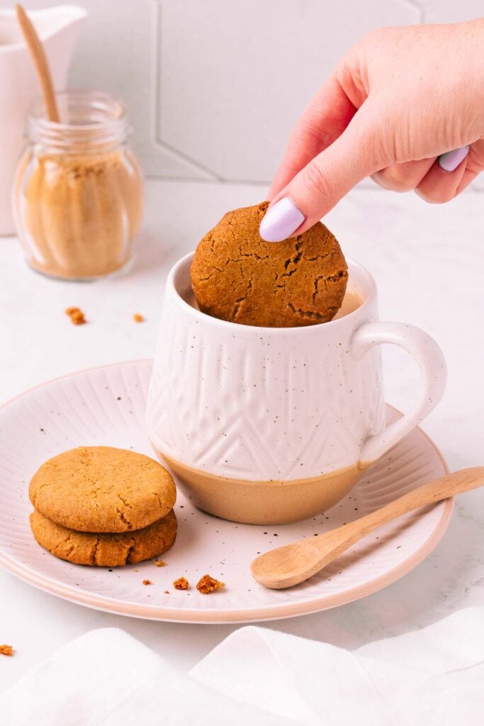 A hand with light purple painted fingernails dunking a gluten free gingernut biscuit into a patterned white mug full of milky tea, on a matching plate with two more gingernuts and a spoon.