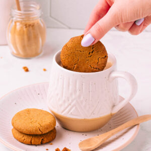 A hand with light purple painted fingernails dunking a gluten free gingernut biscuit into a patterned white mug full of milky tea, on a matching plate with two more gingernuts and a spoon.