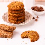 A bitten gluten-free Weet-Bix biscuit on a grey marble surface, with a stack of biscuits, two Weet-bix and a small bowl of chocolate chips in the background.