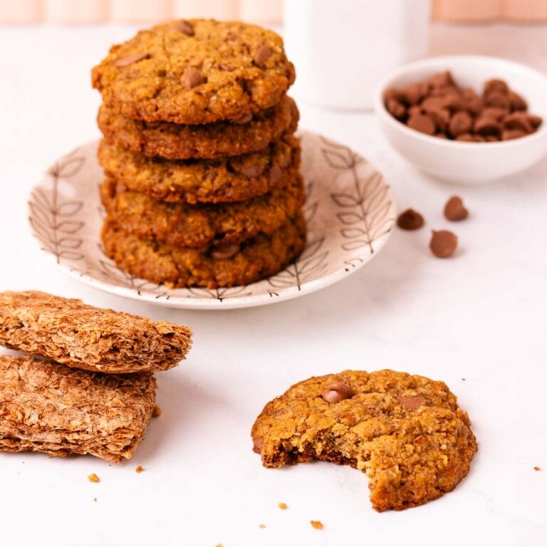 A bitten gluten-free Weet-Bix biscuit on a grey marble surface, with a stack of biscuits, two Weet-bix and a small bowl of chocolate chips in the background.