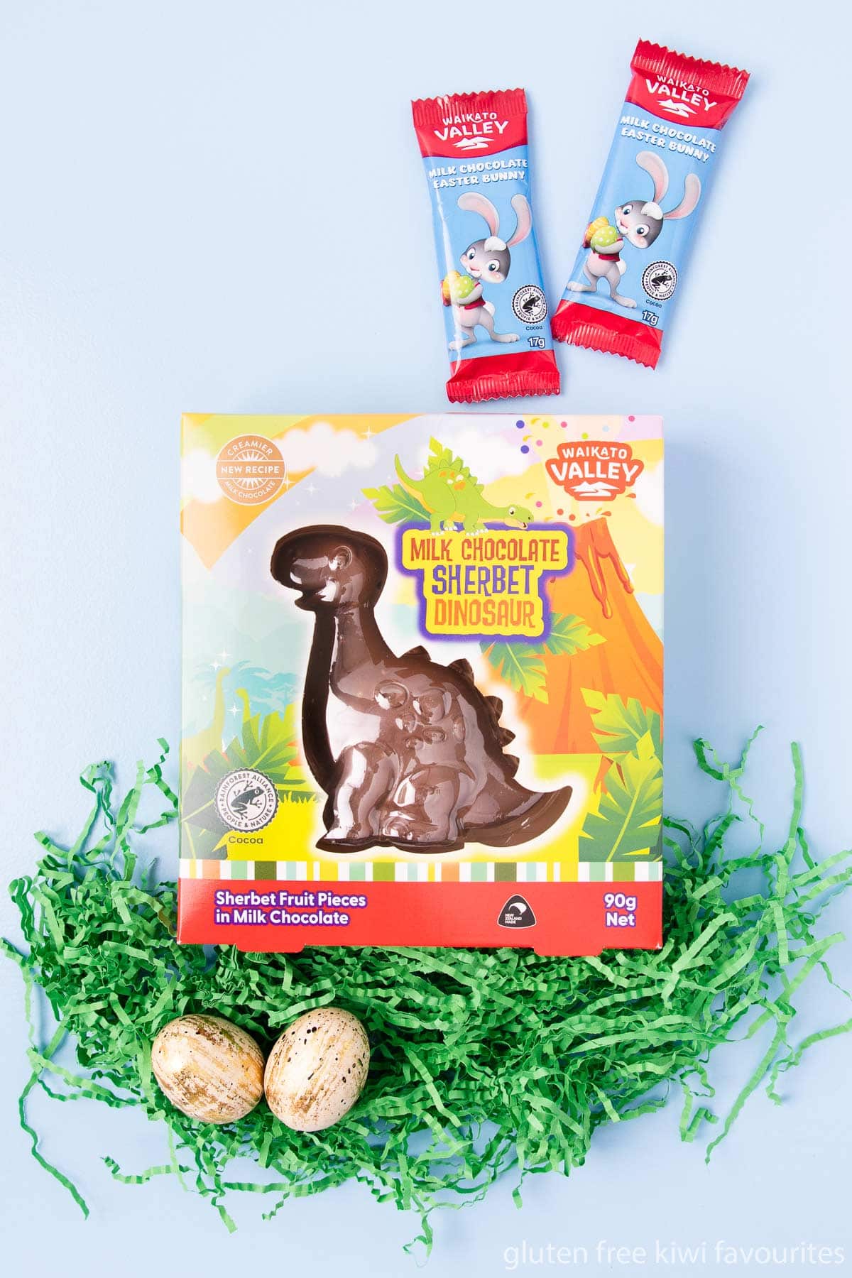 A Waikato Valley brand milk chocolate sherbet dinosaur and two little milk chocolate Easter bunny bars, on a blue background with green shredded paper.