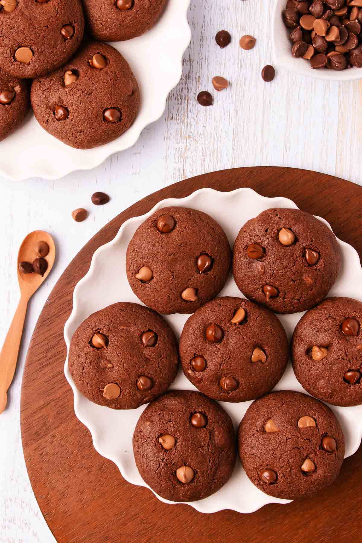 A white scalloped-edge ceramic plate with 7 gluten free chocolate biscuits, on a dark brown wooden round placemat, with a wooden spoon and scattered chocolate chips.