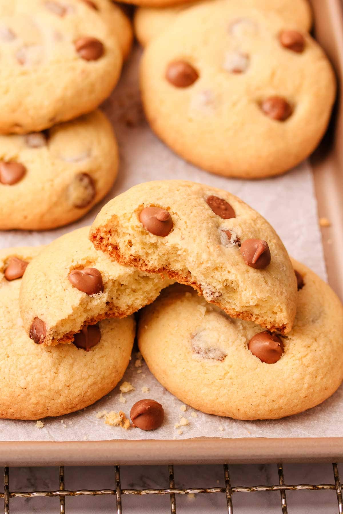 Close-up of two halves of a broken gf chocolate chip biscuit stacked on top of other biscuits, on a baking paper lined rose-gold metal baking tray.