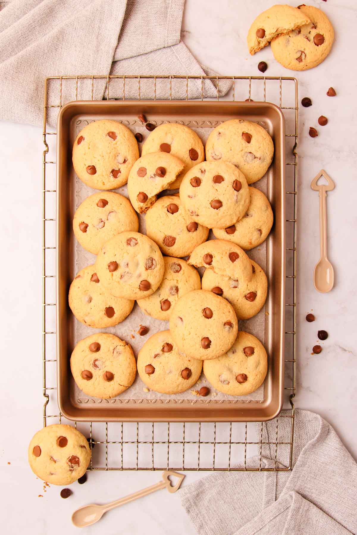 Overhead image of a tray of chocolate chip biscuits on a rustic metal cooking rack, with scattered chocolate chips and crumbs, on a grey marble background.