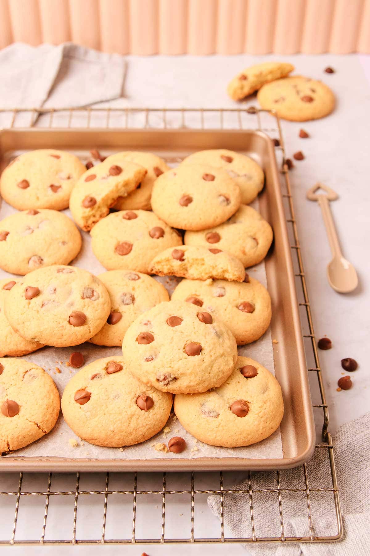 Chocolate chip biscuits scattered haphazardly on a rose gold metal baking tray, which is resting on a rustic metal baking tray, with a cream-coloured ceramic spoon, and scattered chocolate chips.