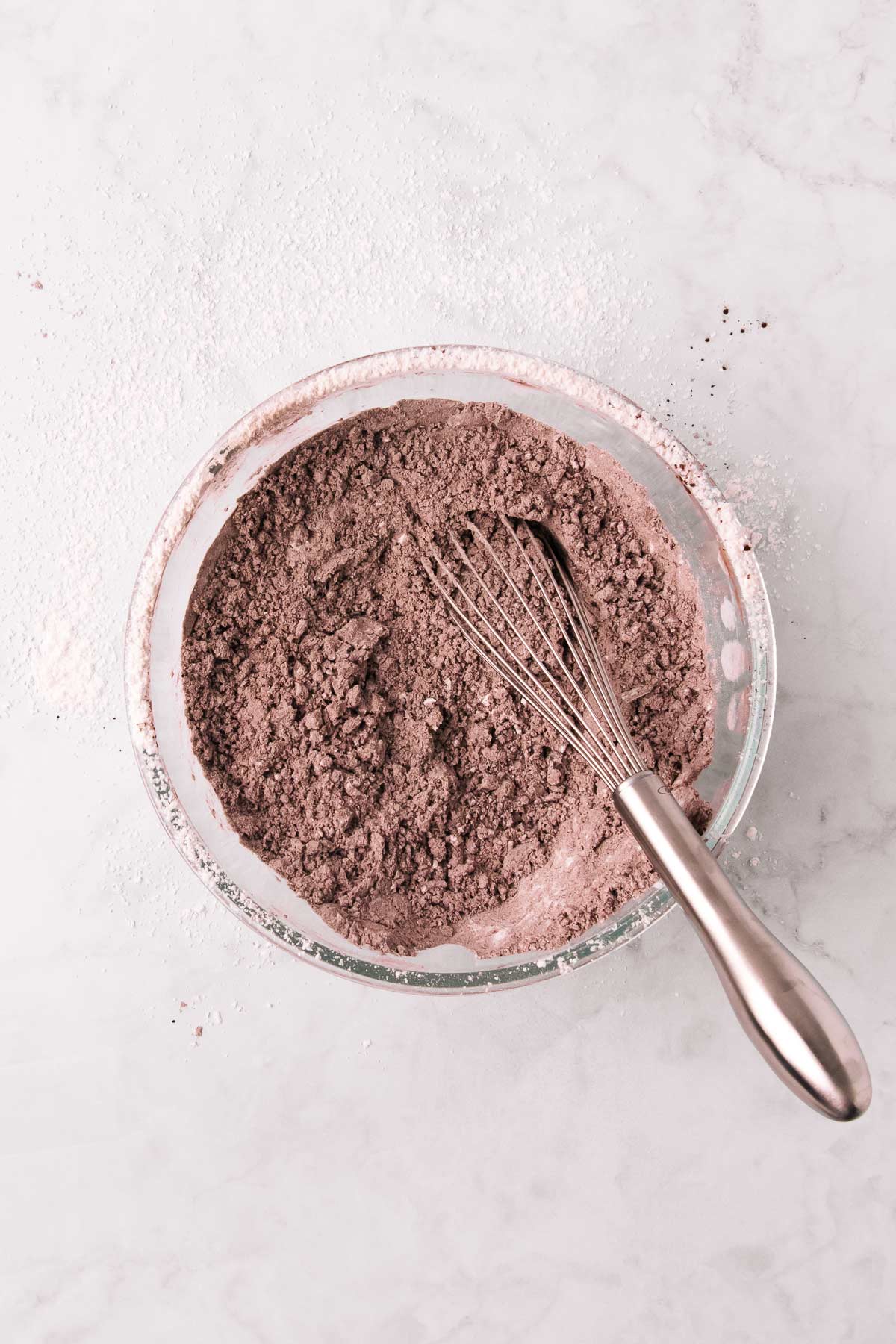 The dry ingredients in a bowl with a small metal whisk, the person who whisked it was messy and there is flour and cocoa all over the sides of the bowl and all over the counter.