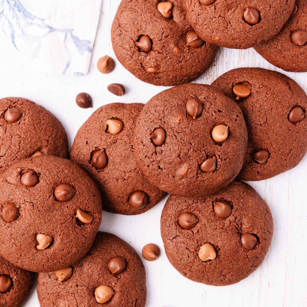 Scattered gluten free chocolate biscuits on a pale blue-washed wooden background, with a blue and white linen napkin in the top left corner.