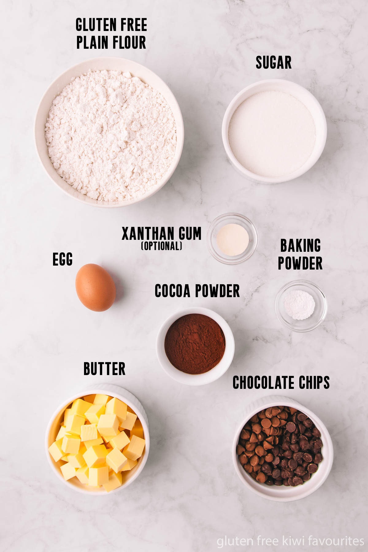 The gluten free chocolate biscuit ingredients - gluten free flour, sugar, baking powder, baking soda, xanthan gum, cocoa powder, egg, butter and chocolate chips - in bowls on a grey marble background.