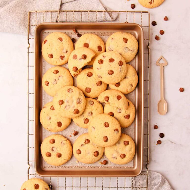 Overhead image of a tray of gluten free chocolate chip biscuits on a rustic metal cooking rack, with scattered chocolate chips and crumbs, on a grey marble background.