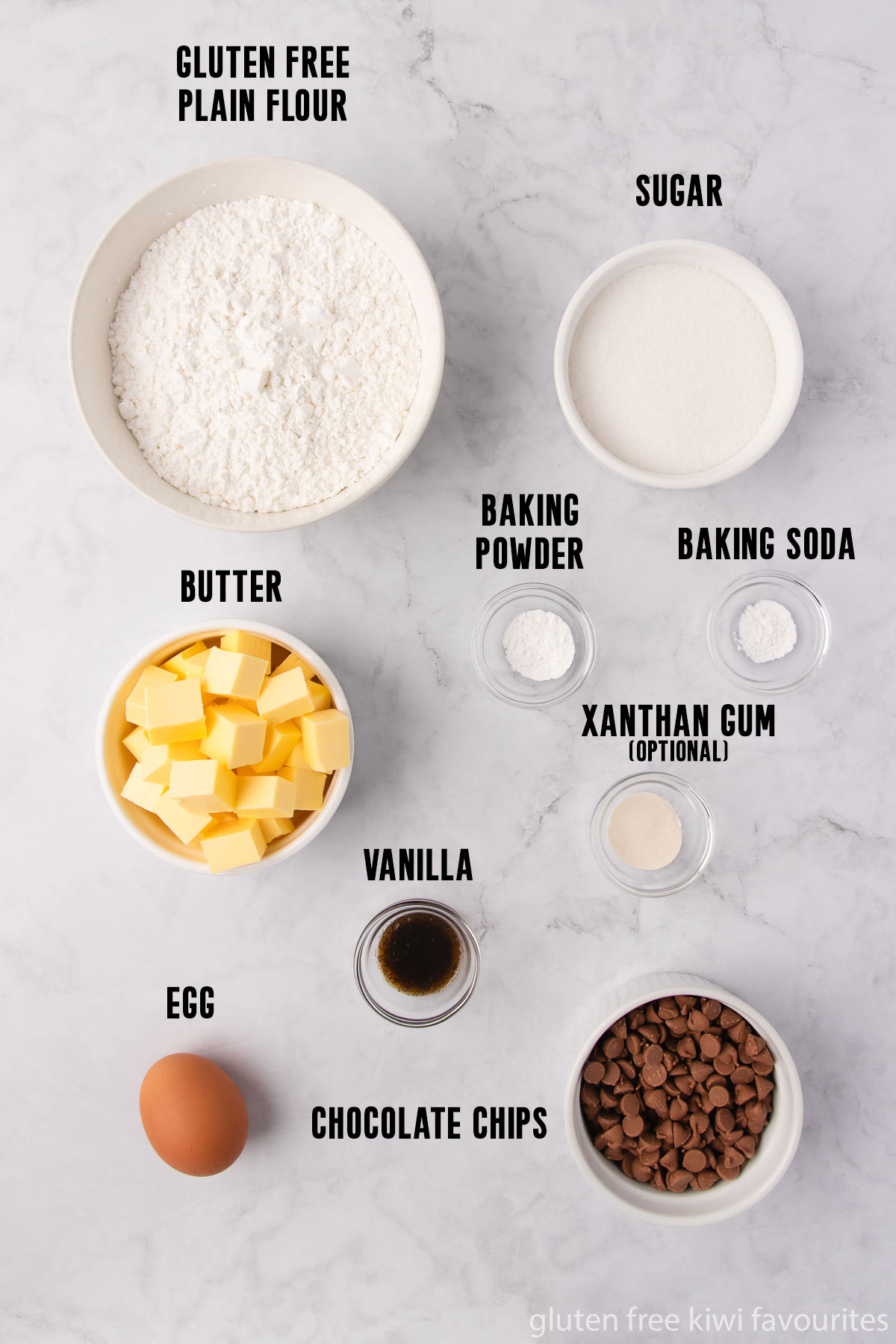 The gluten free chocolate chip biscuit ingredients - gluten free flour, sugar, baking powder, baking soda, xanthan gum, vanilla, egg, butter and chocolate chips - in bowls on a grey marble background.