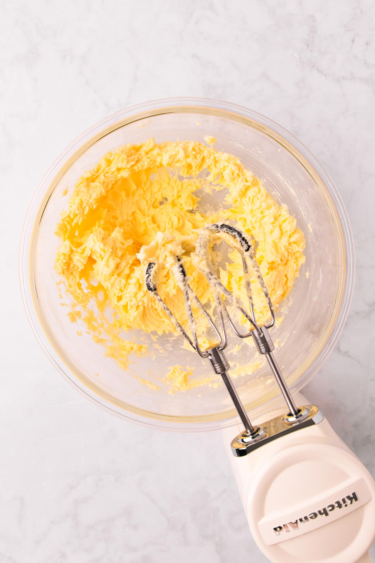Creamed butter, sugar, vanilla and egg in a large glass bowl, with a white Kitchenaid handheld electric mixer.