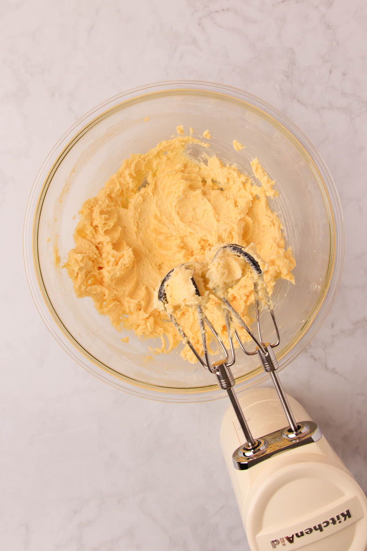 The creamed butter, sugar, vanilla and egg mixture in a large glass bowl, with a white Kitchenaid handheld electric mixer.