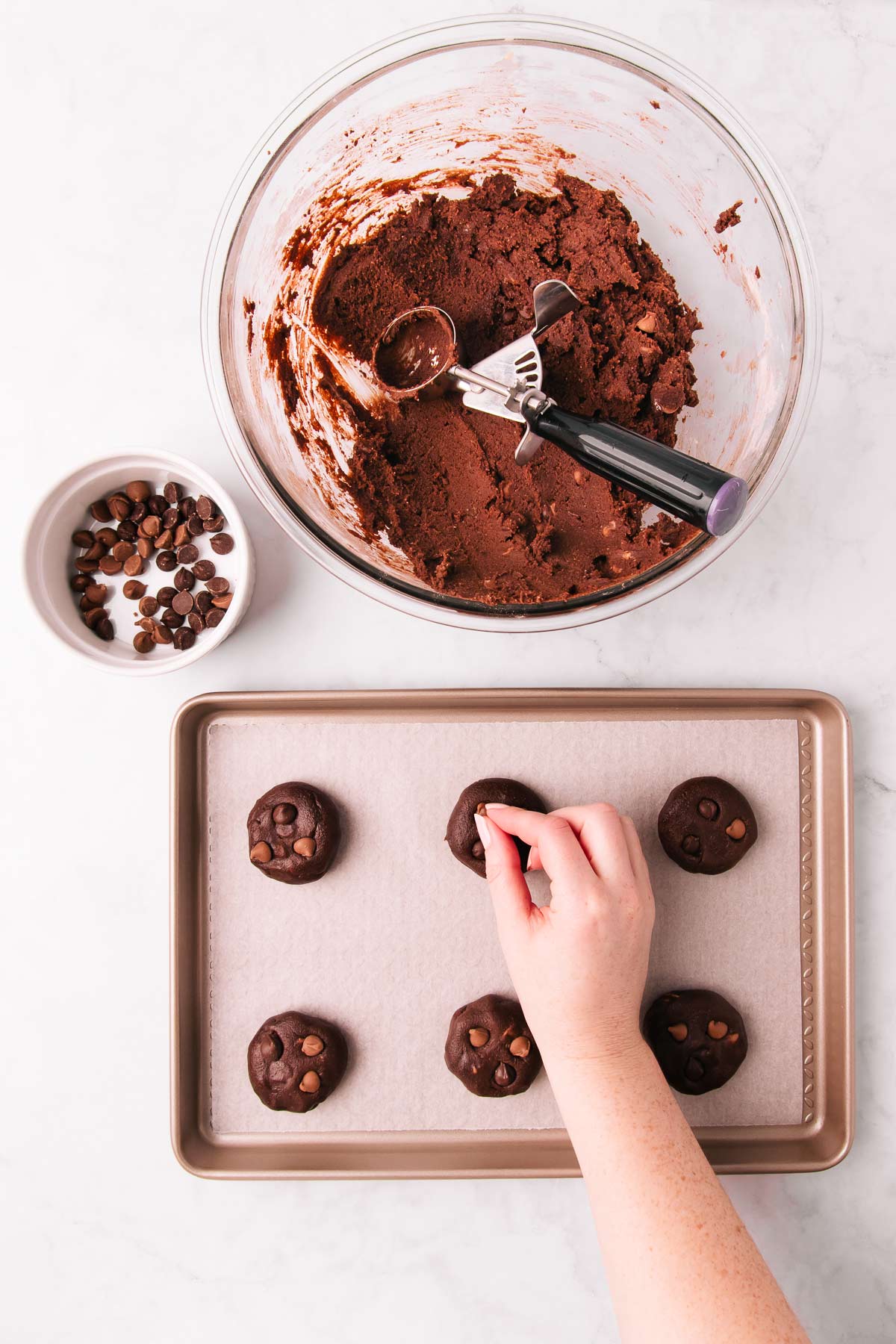 The bowl of biscuit dough, and a small baking tray with balls of dough on it. A hand is pressing chocolate chips into the tops of the balls of dough.