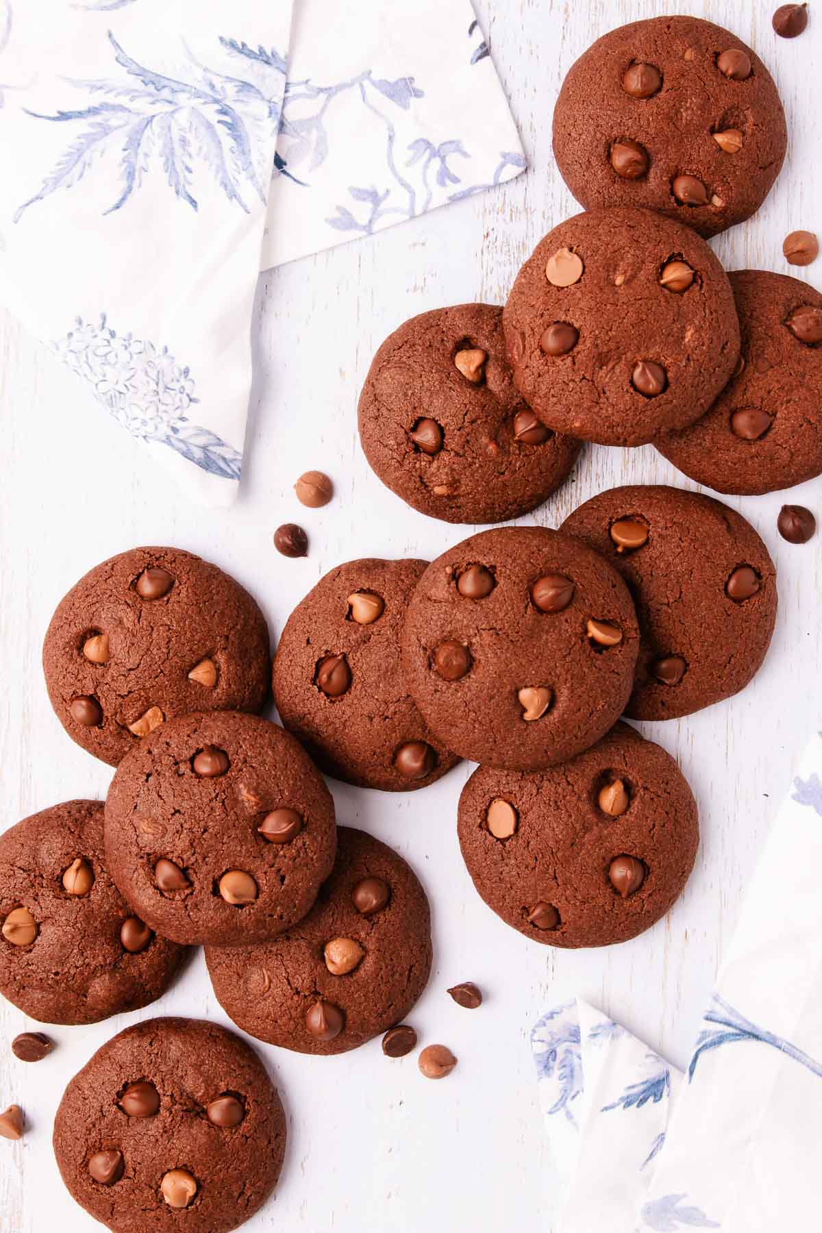 Scattered gluten free chocolate biscuits on a pale blue-washed wooden background, with a blue and white linen napkin in the top left and bottom right corners.
