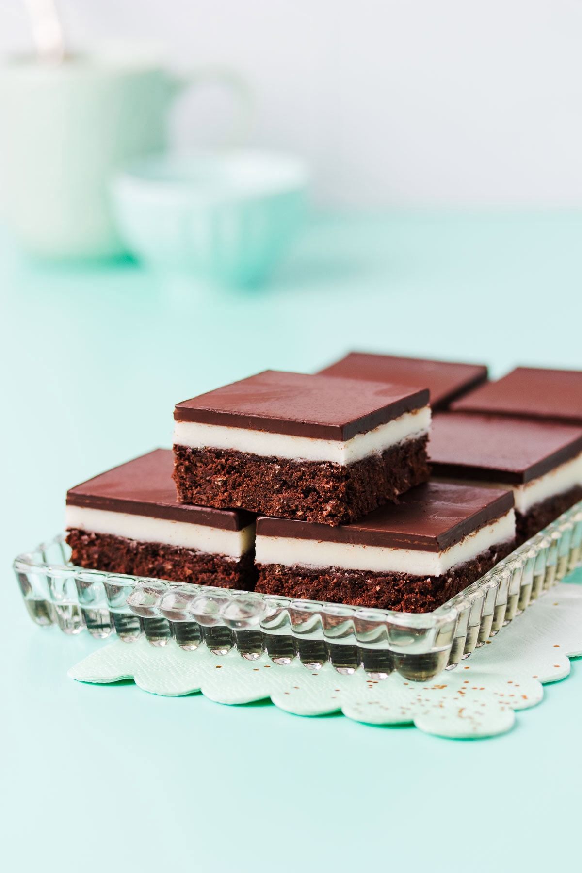 Pieces of gluten-free peppermint slice on a rectangular glass plate, with a mint-green paper napkin on a mint green background.