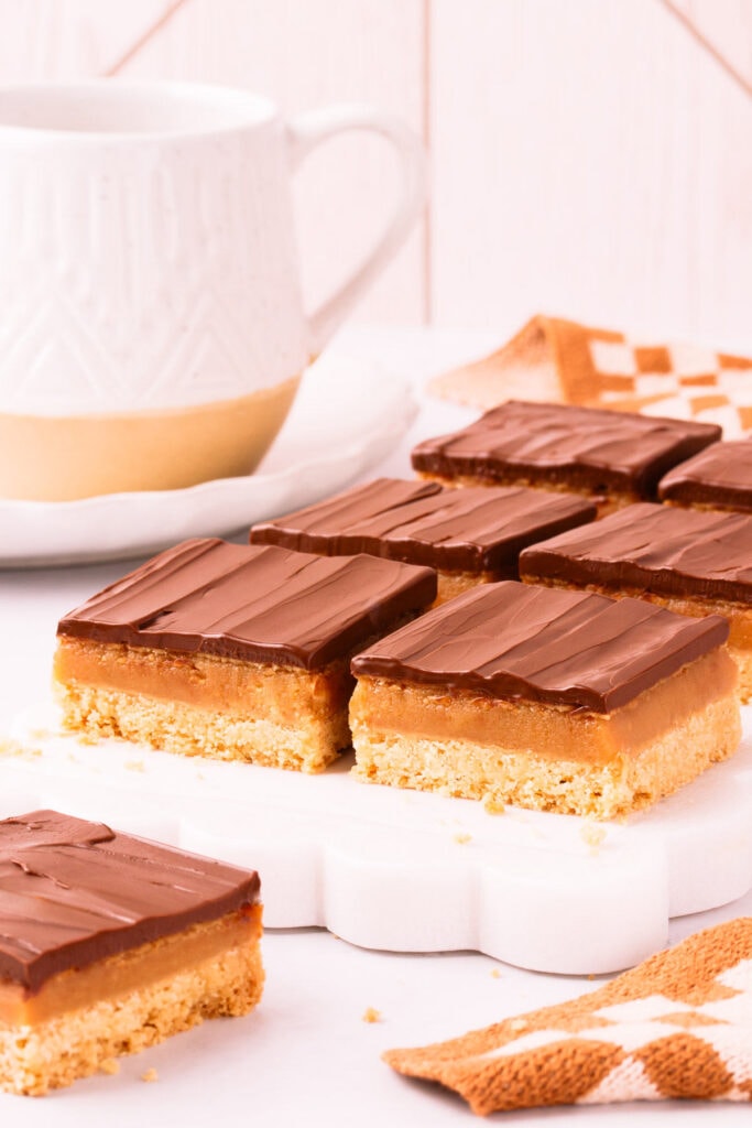 Six square pieces of gluten free caramel slice on a white marble slab with scalloped edges, on a light grey background with a speckled white mug of tea in the background.