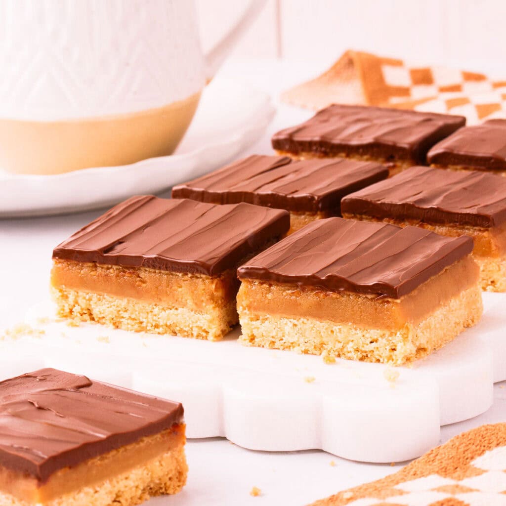 Six square pieces of gluten free caramel slice on a white marble slab with scalloped edges, on a light grey background with a speckled white mug of tea in the background.