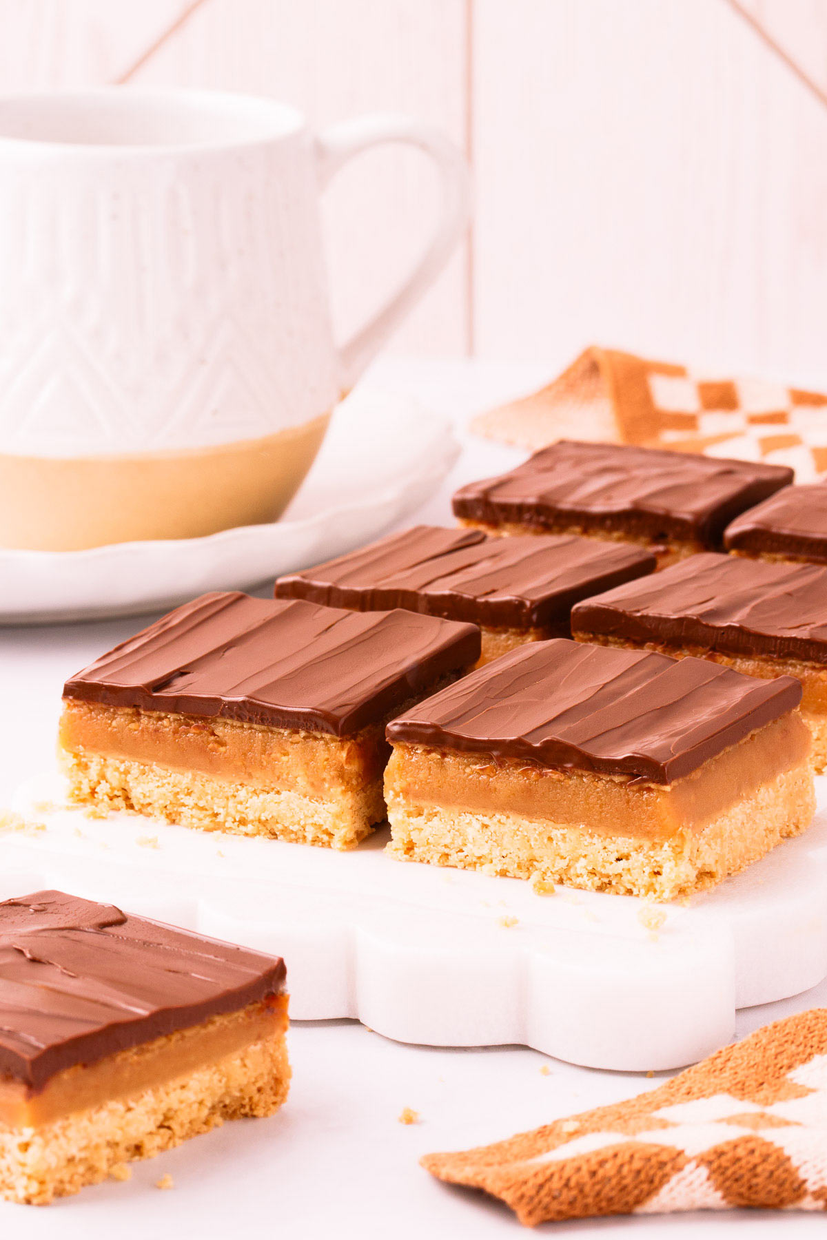 Six square pieces of gluten free caramel slice on a white marble slab with scalloped edges, on a light grey background with a speckled white mug of tea in the background.