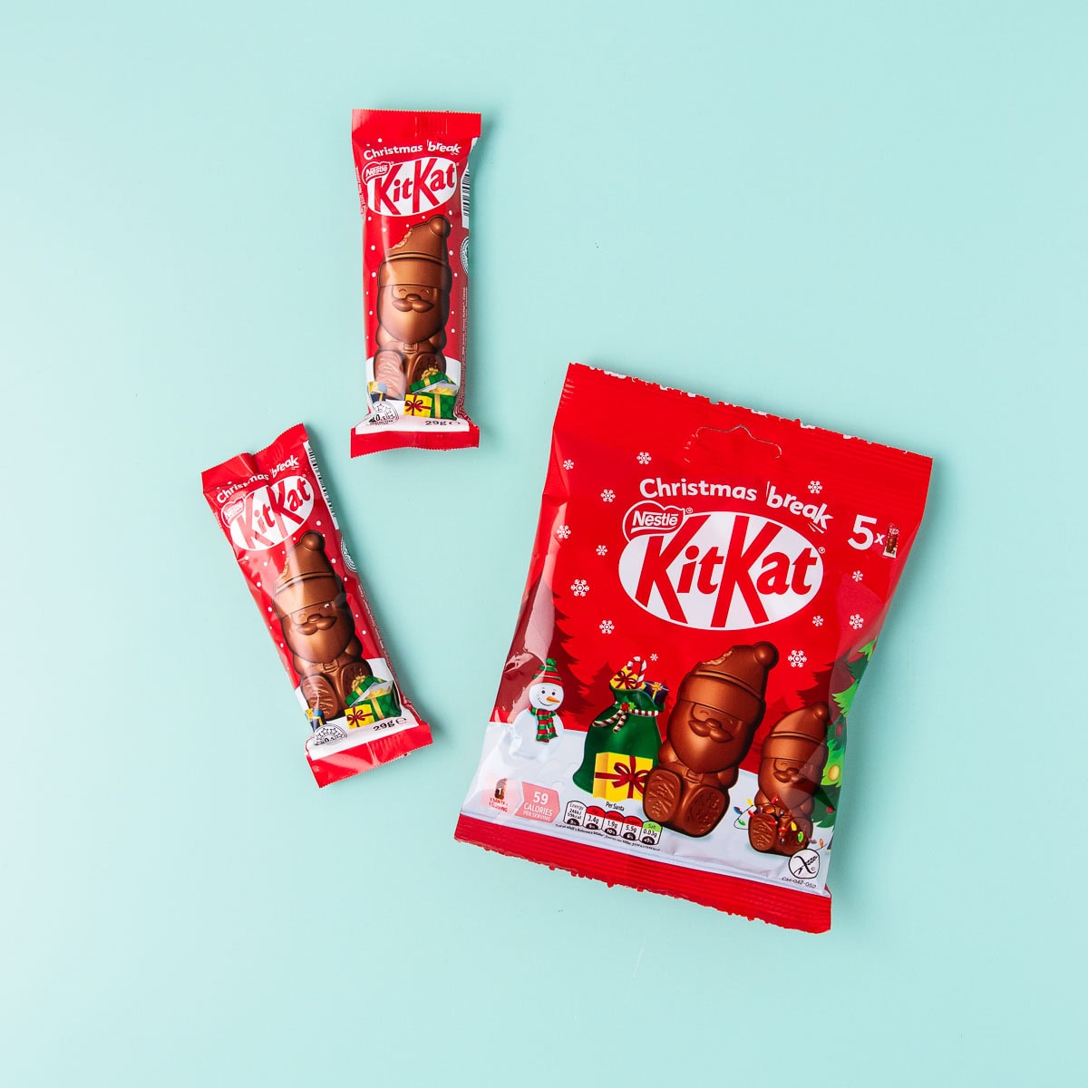 A red package of KitKat mini chocolate Santas and two individual KitKat Santas on a mint green background.