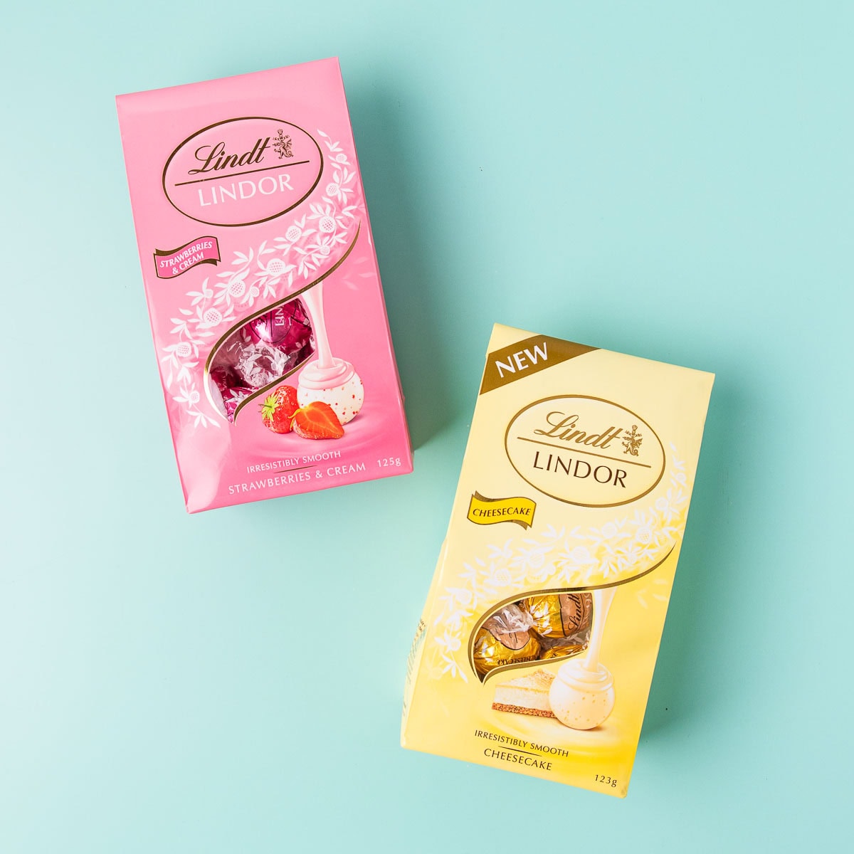 A pink packet of Lindt strawberries and cream truffles, and yellow packet of cheesecake truffles, on a mint green background.