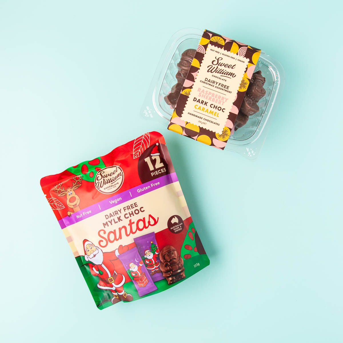 A packet of Sweet William dairy free mylk choc Santas and a container of Sweet William dairy free Christmas assortment chocolates on a mint green background.